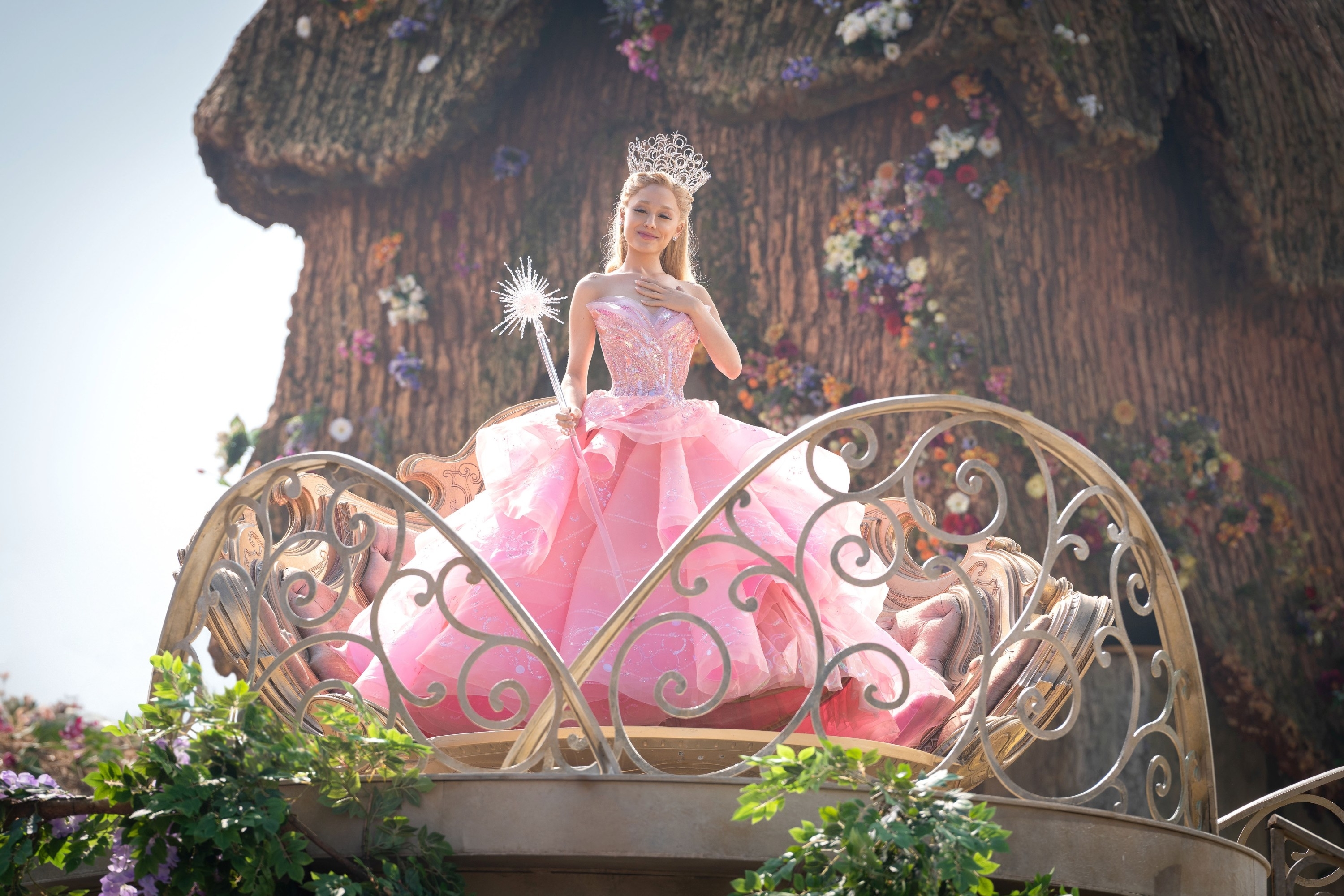 Glinda with a tiara and wand, in a voluminous gown, stands on a decorative balcony set against a fantasy backdrop