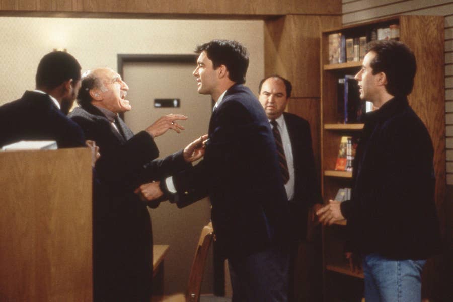 Scene from a TV show with four men in a tense confrontation inside a bookstore, featuring actors in business and casual attire