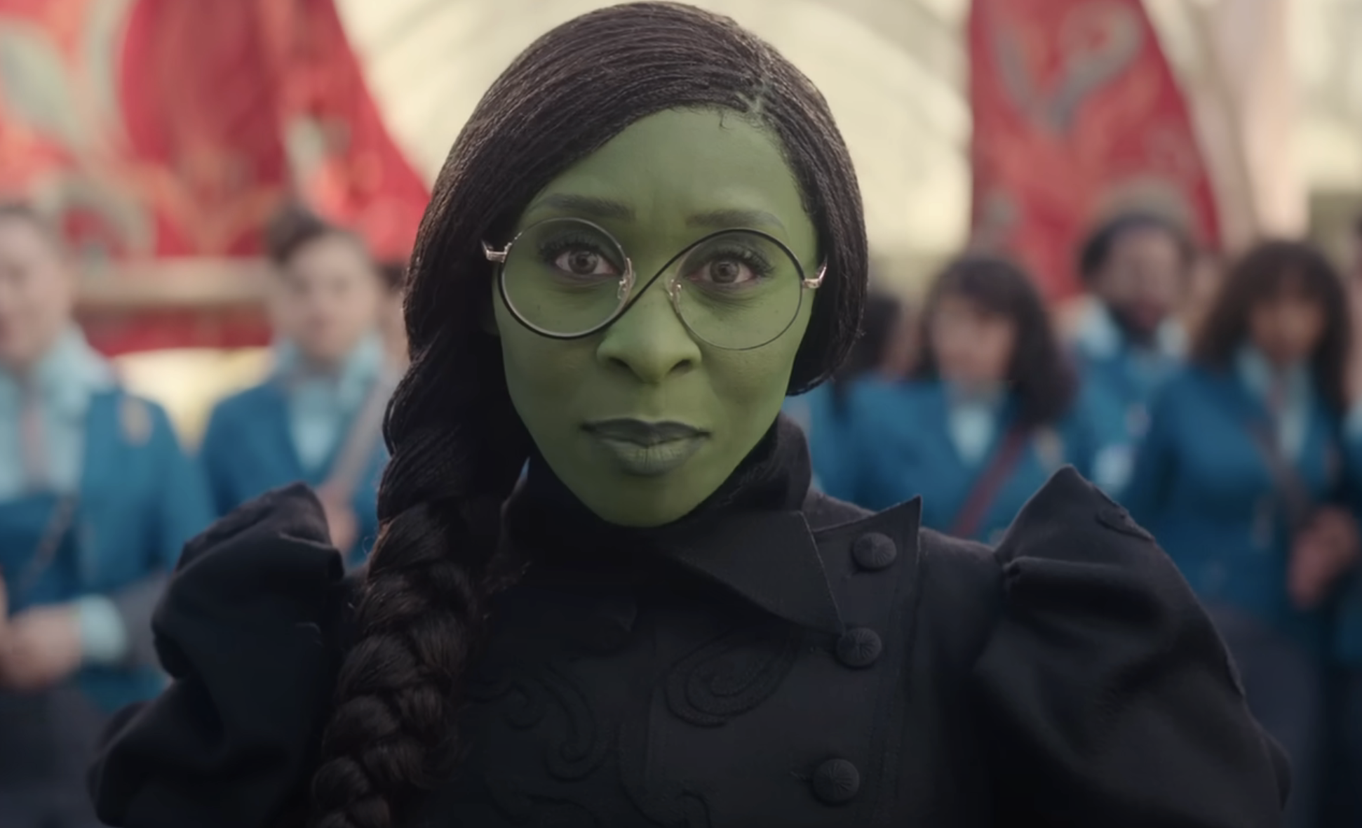 Elphaba with microbraids in a long braid, wearing a dark dress with puffy sleeves and round, unique glasses
