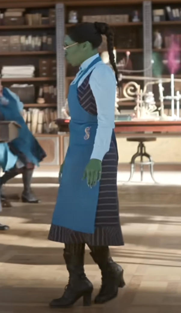 Elphaba wearing a long blue apron over a striped outfit with tall boots with hair in a long braid