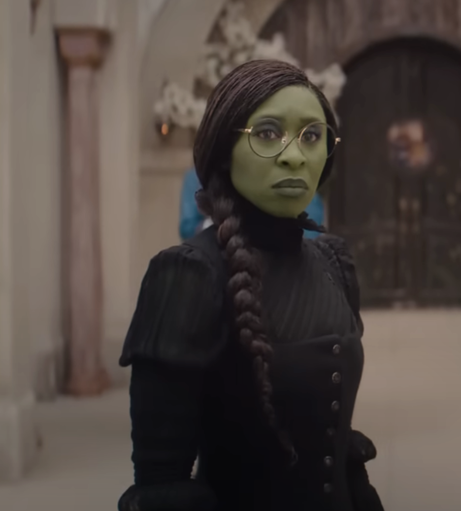 Elphaba in dark dress with long braided hair, standing in an ornate hall