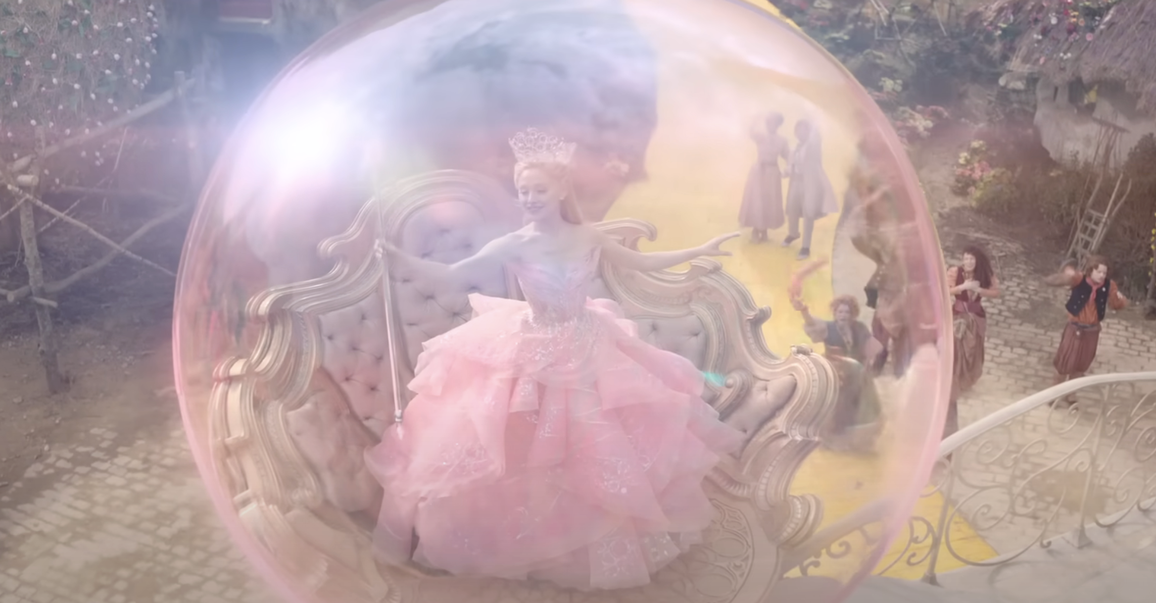 Glinda in a gown and crown inside a large bubble, seated on an ornate chair, surrounded by onlookers in rustic attire