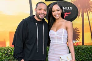 Two people pose on a red carpet; one in a casual black outfit, the other in an elegant light-colored strapless dress holding a clutch. Netflix event backdrop