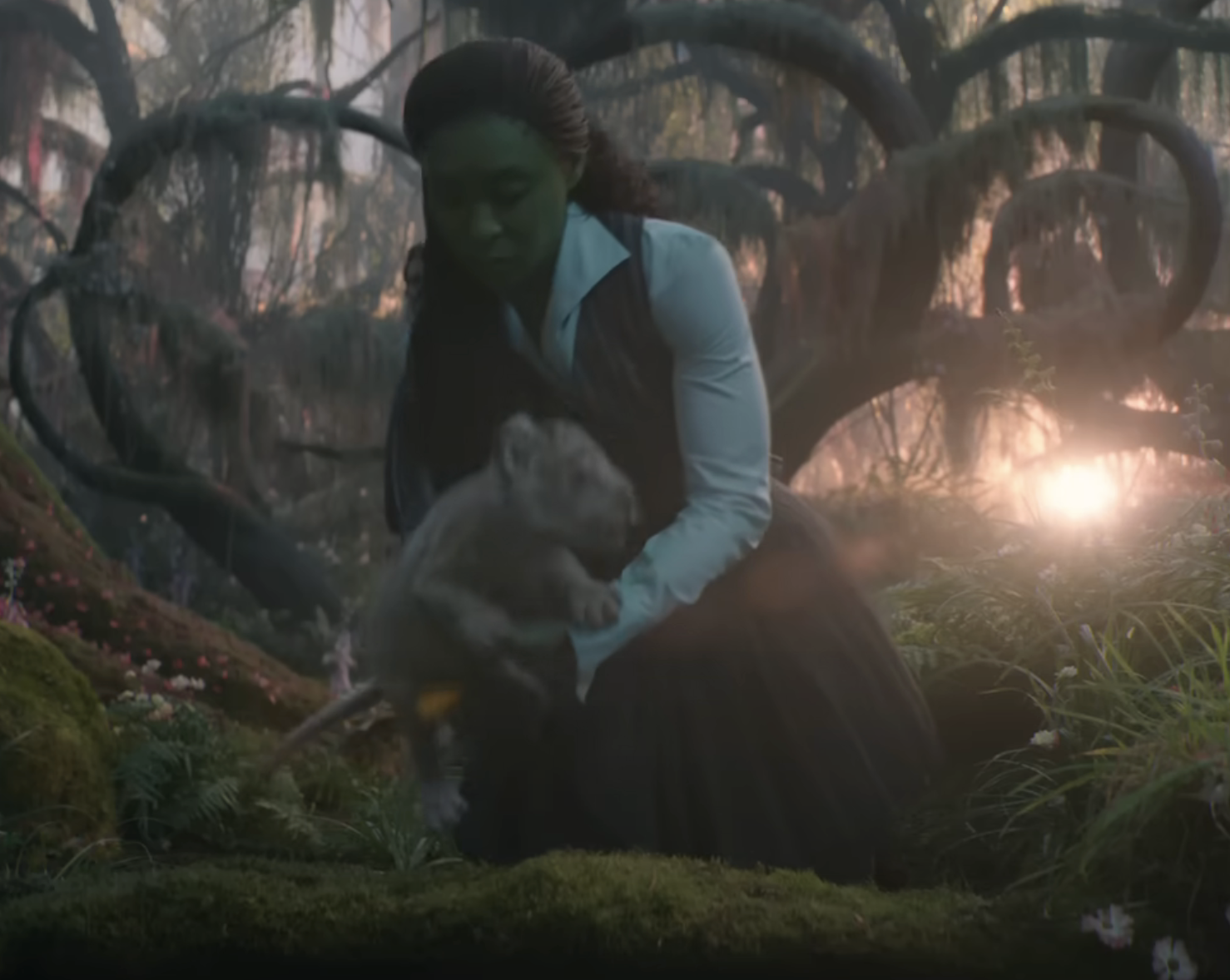Elphaba in a pinafore with hair pulled back sets down a lin cub in a lush forest