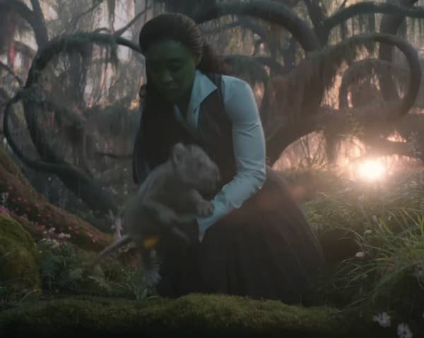 Elphaba in a pinafore with hair pulled back sets down a lin cub in a lush forest
