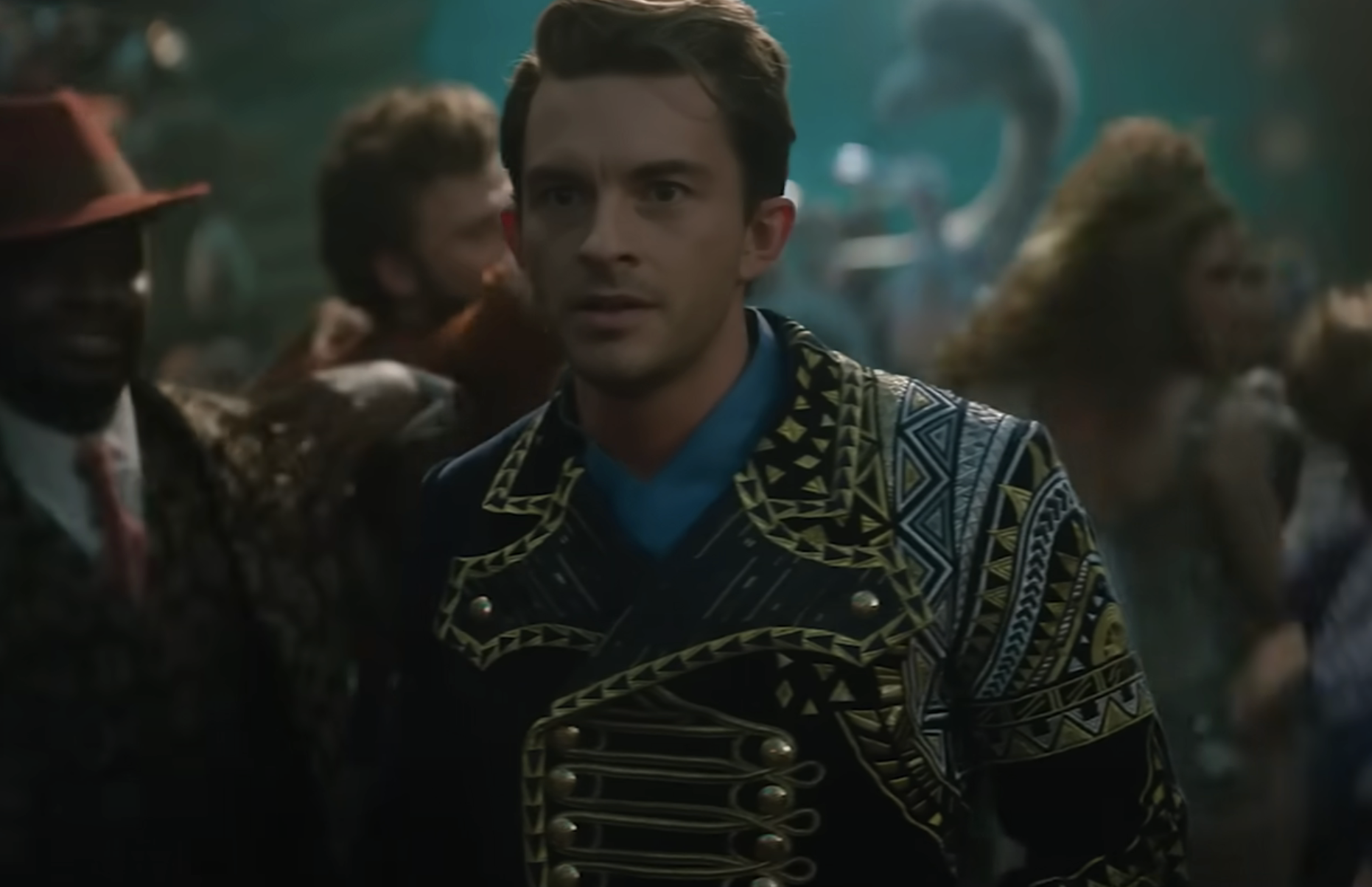 Fiyero wearing an ornate, patterned jacket stands in a crowded, lively setting