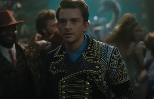Fiyero wearing an ornate, patterned jacket stands in a crowded, lively setting