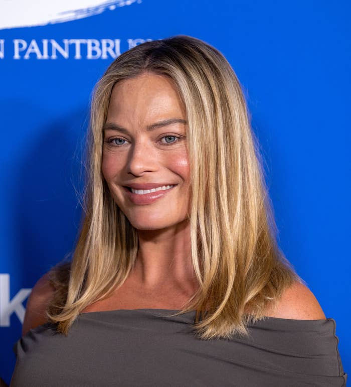 Margot Robbie with long hair wearing an off-shoulder dress, smiling, standing against a blue background