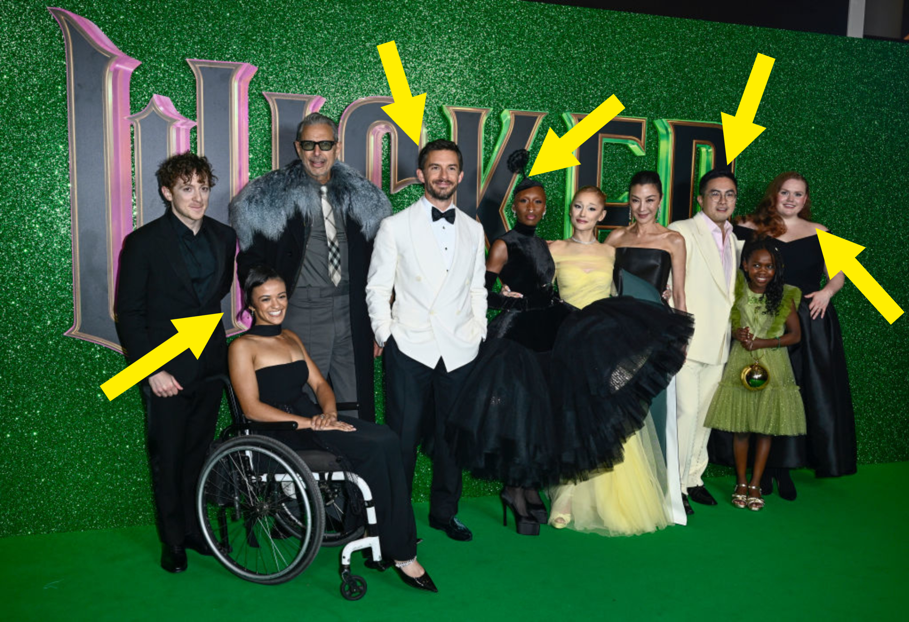 Group of people posing on a green-themed event, featuring formal and elegant attire. One person is in a wheelchair, and dresses include black and yellow gowns