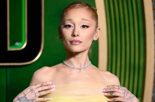 Person posing on red carpet, wearing an off-shoulder gown and choker necklace, with tattoos visible on hands