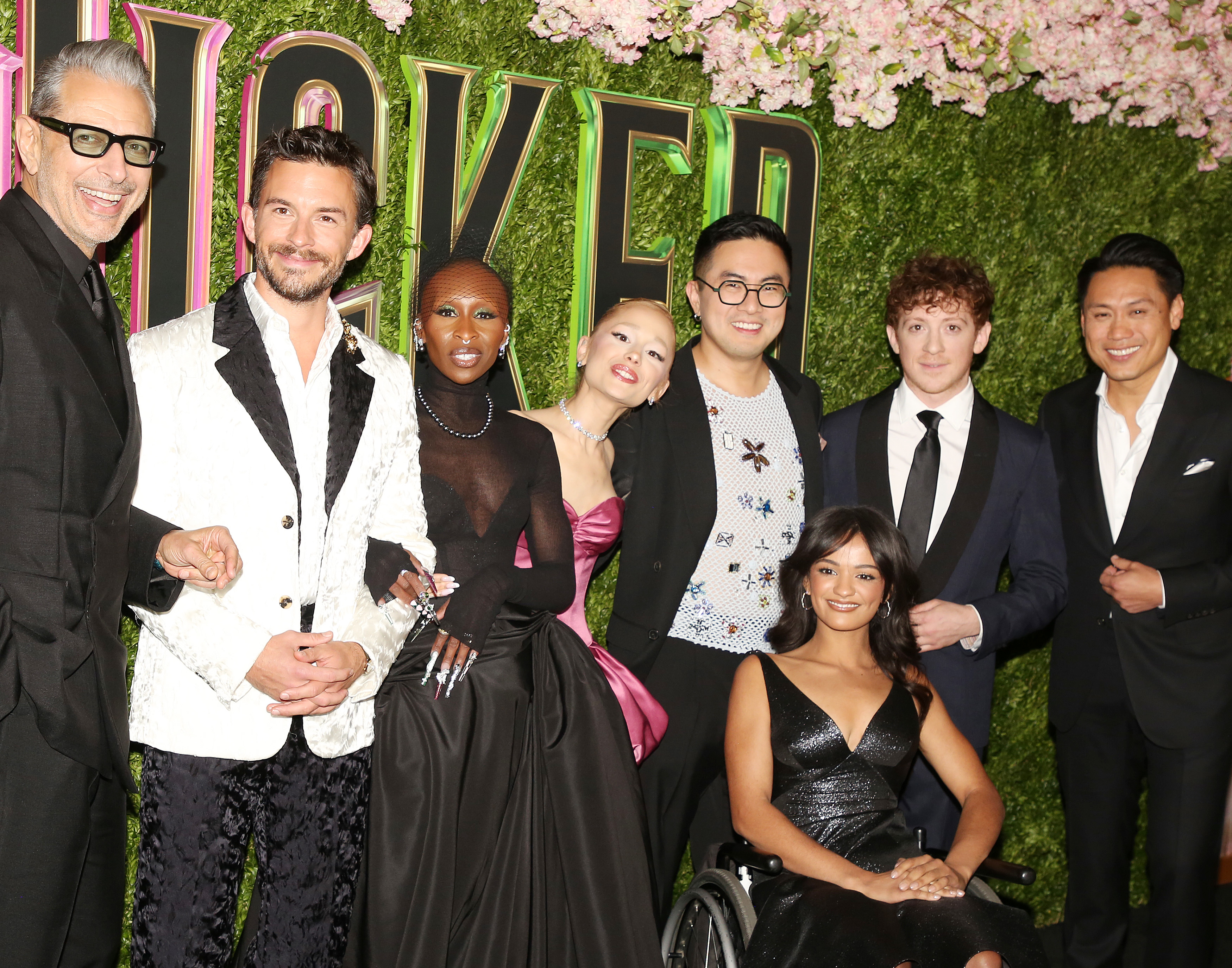 Group of actors posing on a red carpet, dressed in elegant evening attire, against a floral backdrop at a theatrical event