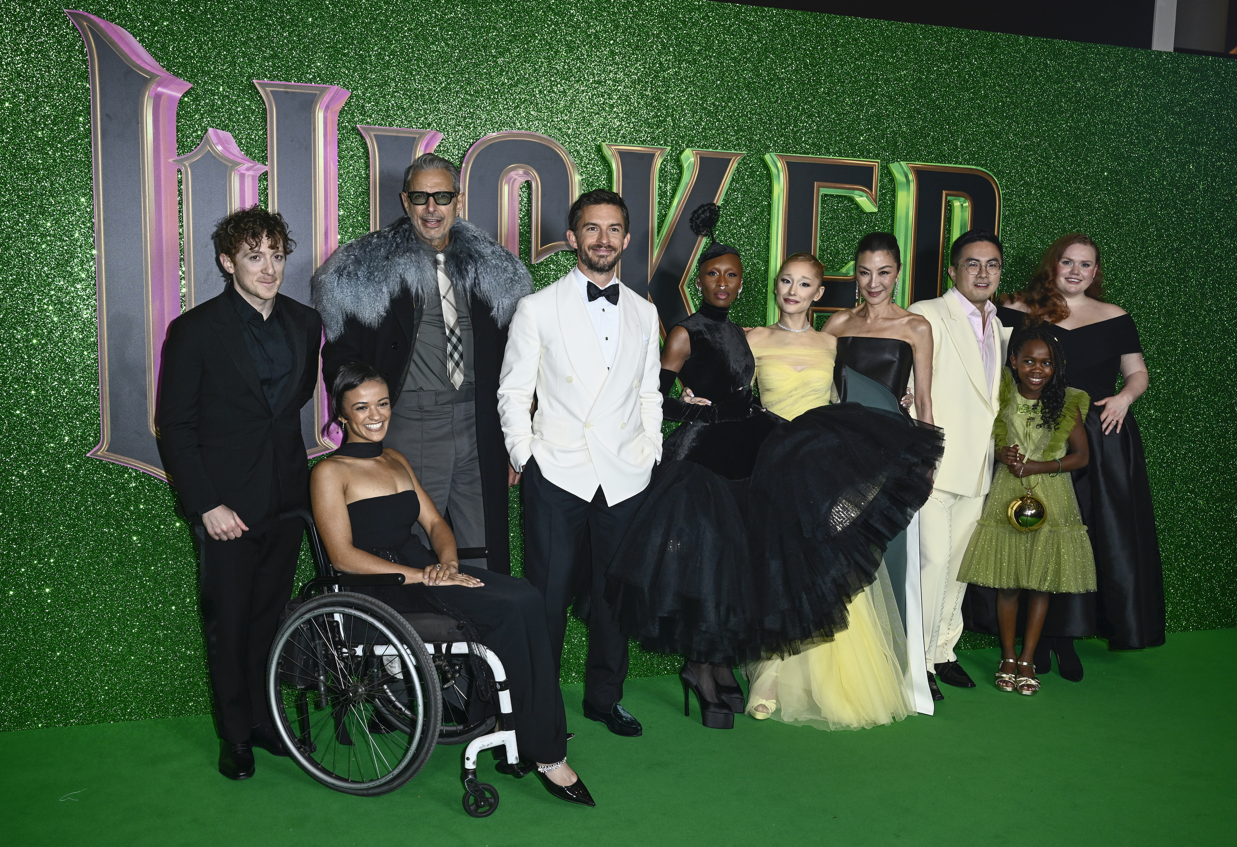 Group of people in formal attire at the &quot;Wicked&quot; event. Some are in black dresses, one in a wheelchair, others in tuxedos, standing on a green carpet