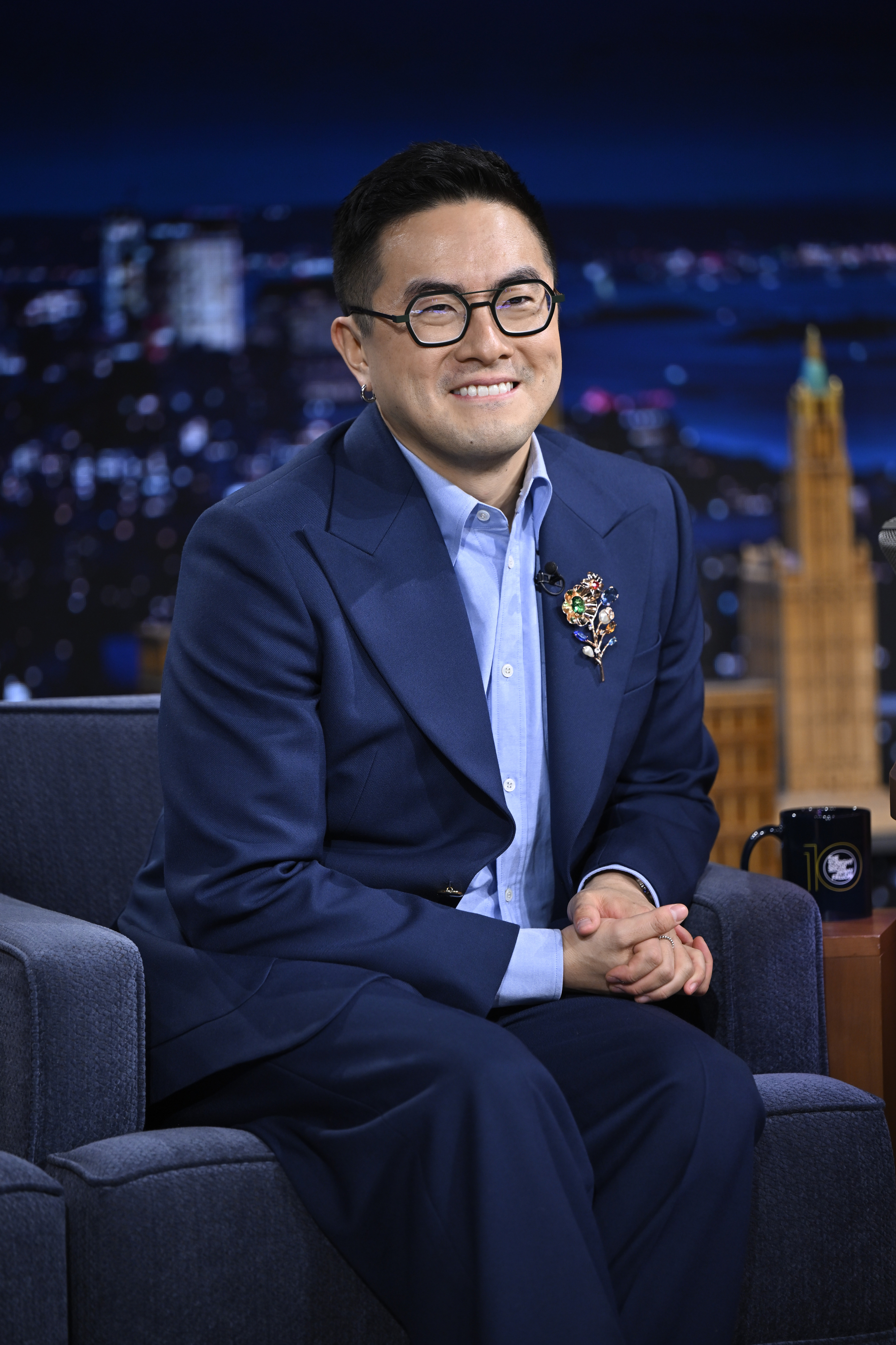 Person in a tailored suit with brooch, sitting on a talk show set, smiling