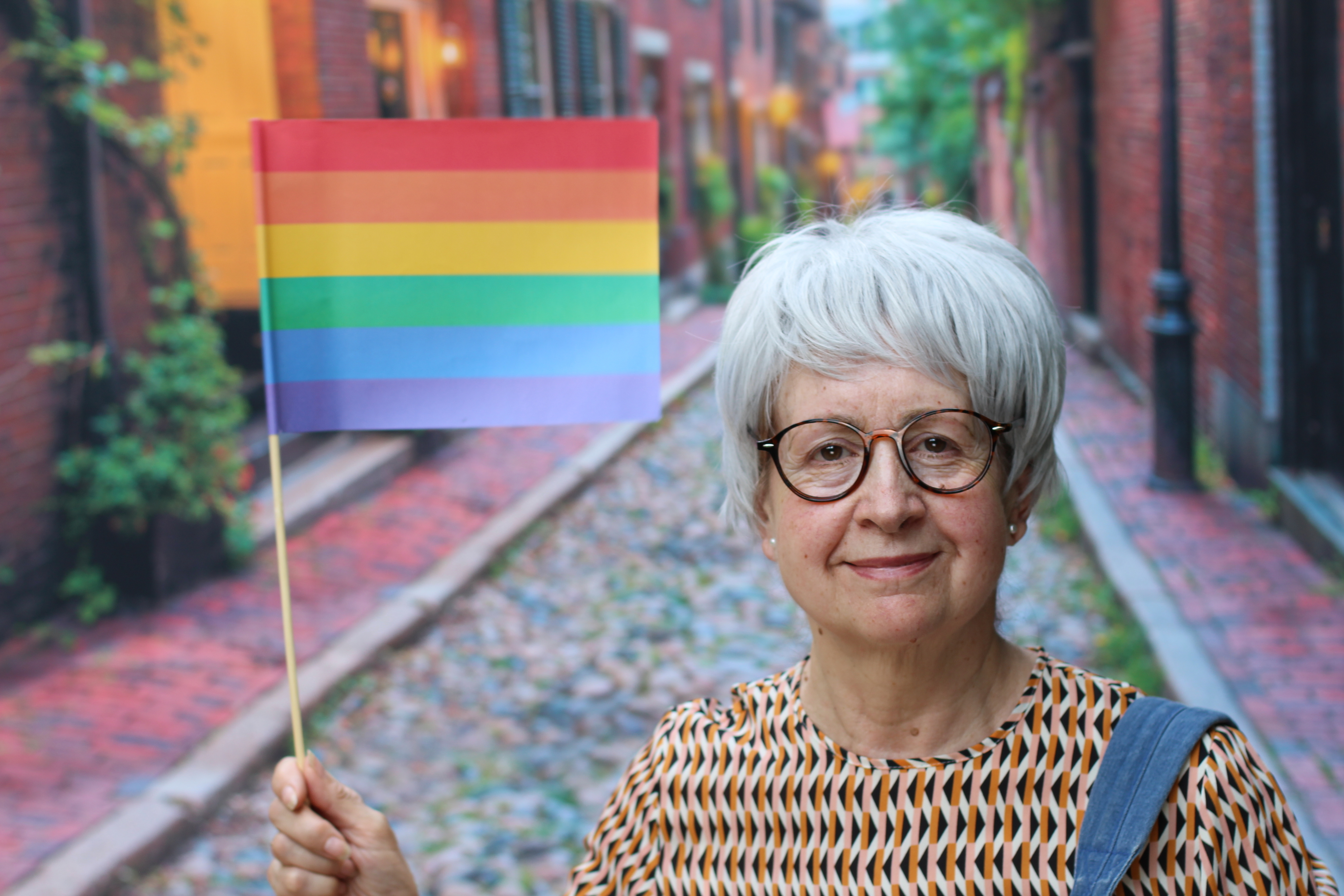 Older woman with glasses holding a rainbow flag, standing on a cobblestone street