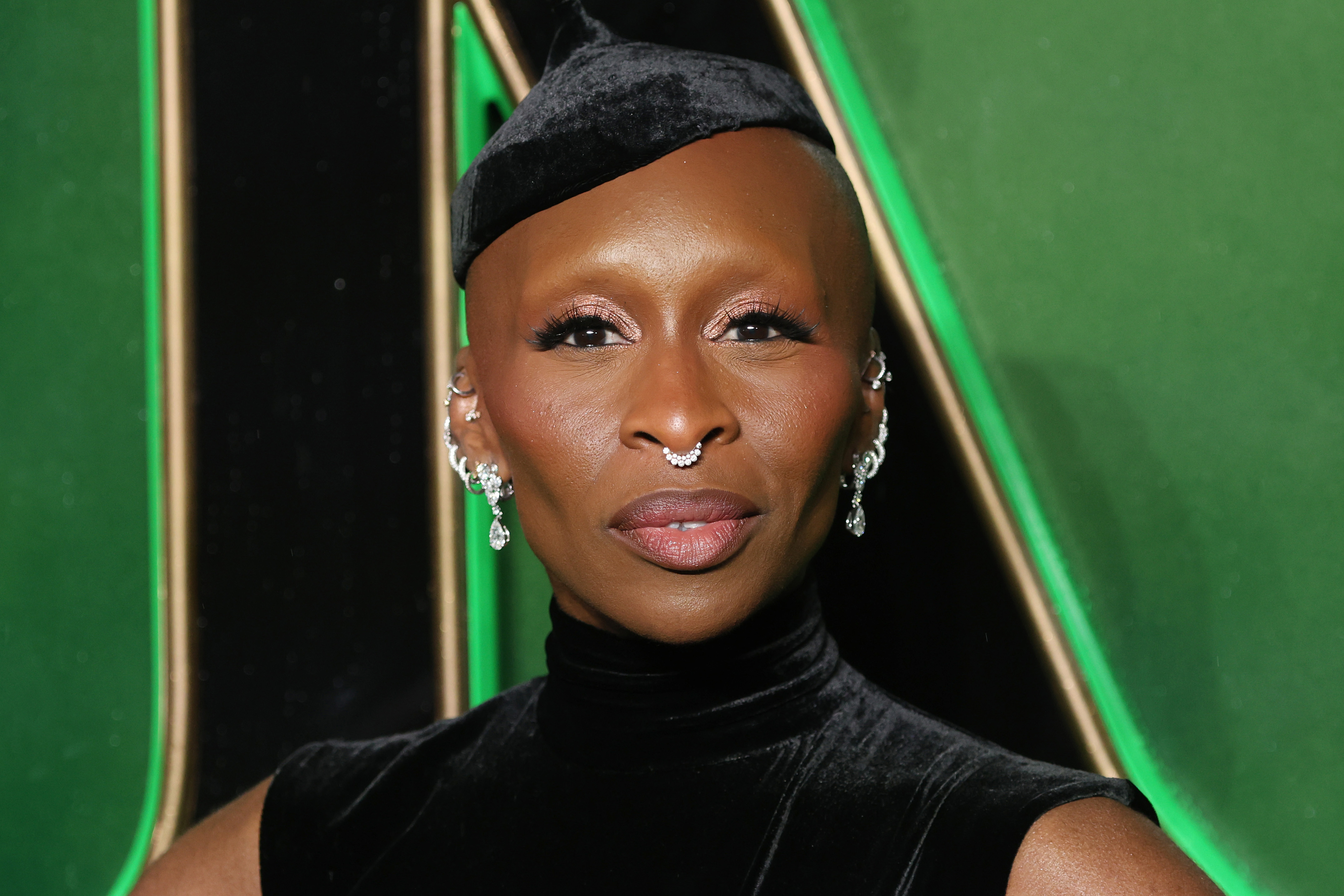 Person wearing a sleek velvet outfit with a stylish beret, adorned with multiple earrings and a nose ring, poses for a photo on a green background