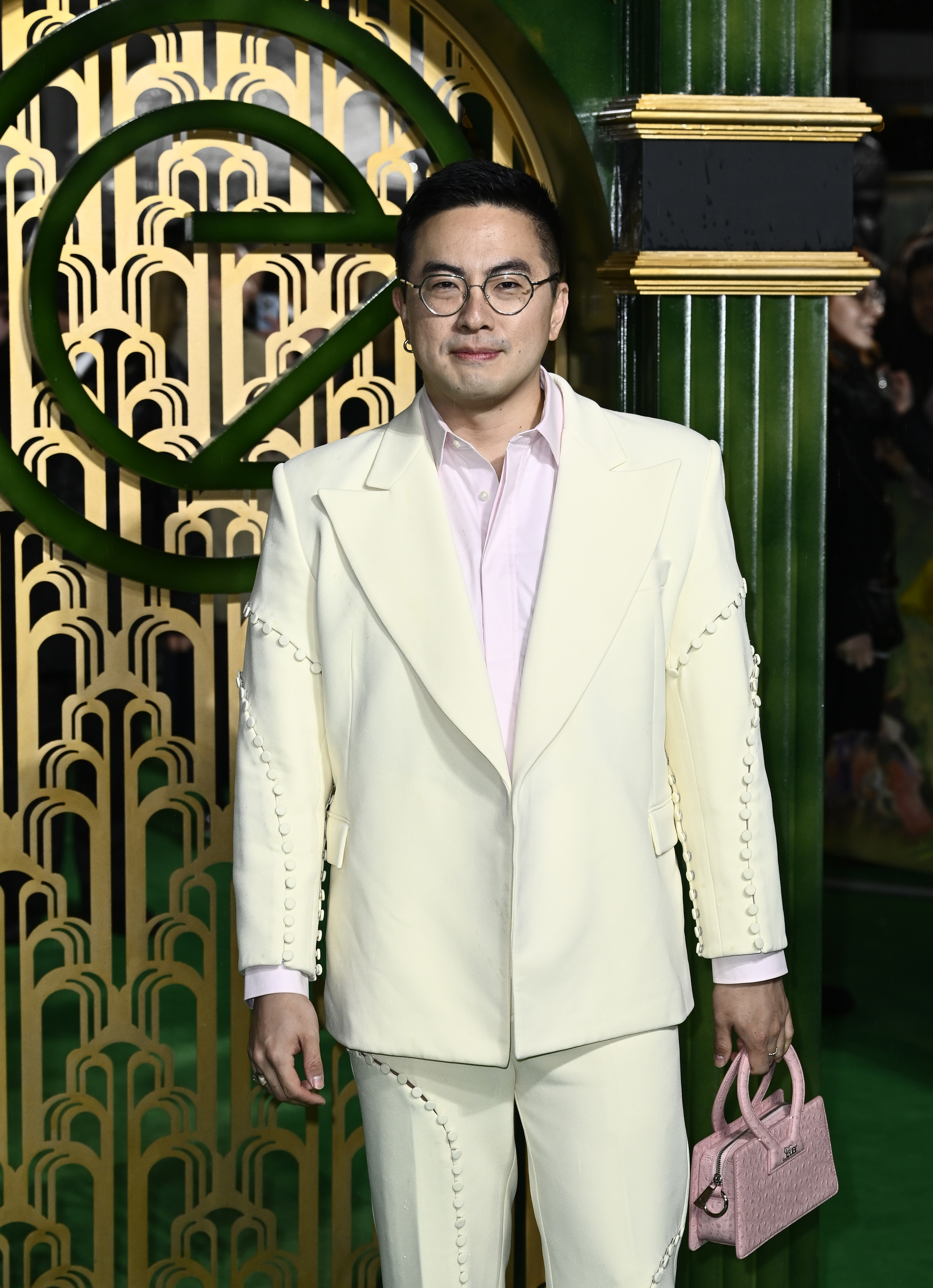 Bowen in a tailored suit with unique stitching detail and shirt, holding a small handbag