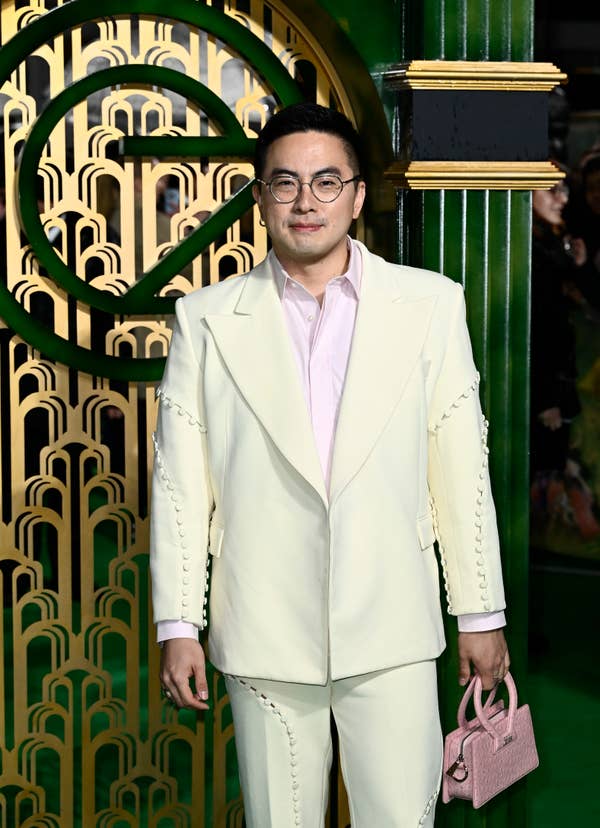 Bowen in a tailored suit with unique stitching detail and shirt, holding a small handbag