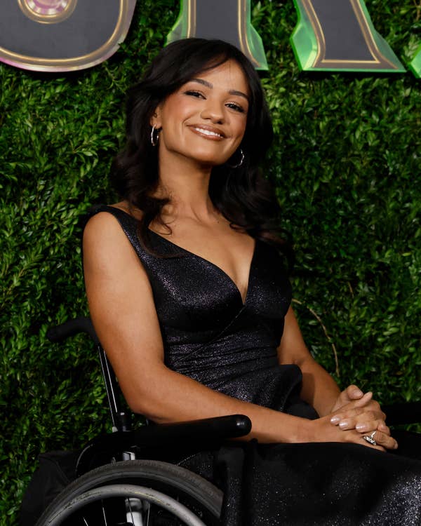 Marissa Bode in a wheelchair wearing a stylish dark dress, smiling at a celebrity event with a greenery backdrop