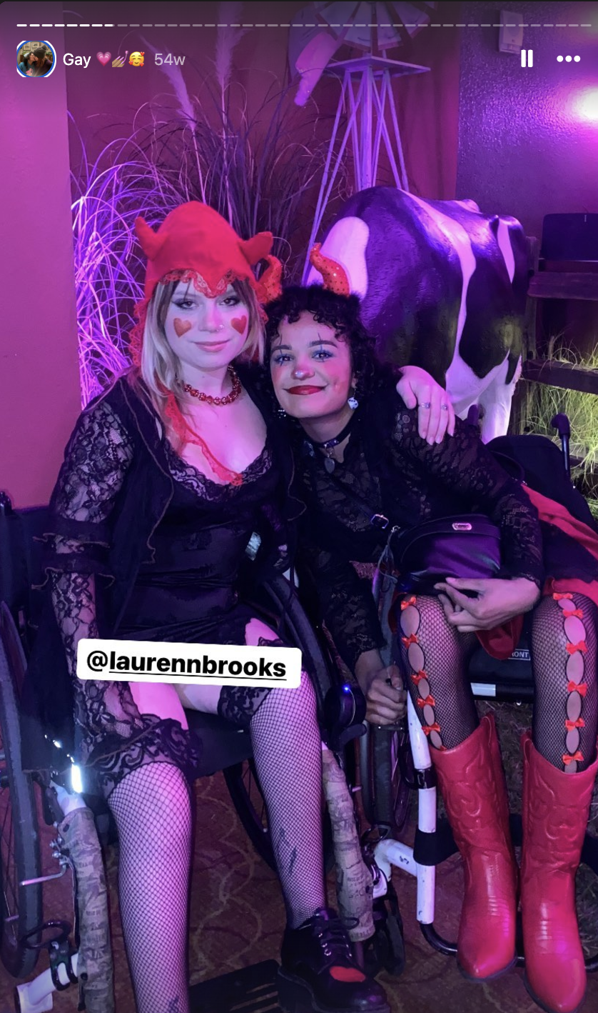 Two people in wheelchairs wearing devil-themed costumes with fishnet tights, lace dresses, and heart face paint at an event