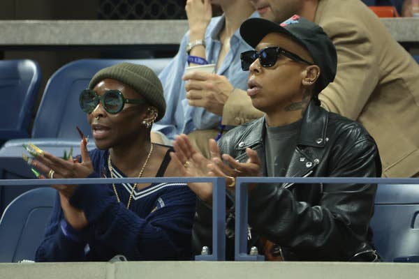 Two people sitting in a stadium, wearing sunglasses and casual outfits, clapping. They appear engaged in the event