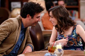 Two people sit at a table in a cozy setting, smiling at each other, with drinks and a candle visible on the table