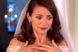 Bride with concerned expression wearing an off-shoulder wedding dress and veil, touching her chest in apparent anxiety