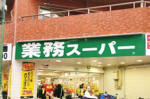 業務スーパーの店舗外観。店の入口近くに顧客がいる。広告看板に油の価格が表示されている。