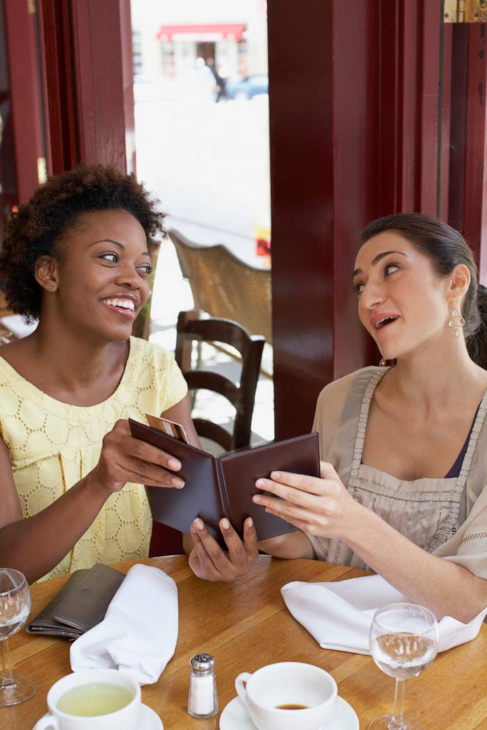 Two women sit at a restaurant table, smiling and holding menus, suggesting a work-related lunch meeting