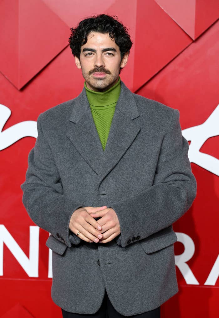 Person in a gray blazer and green turtleneck poses on a red carpet with a geometric backdrop
