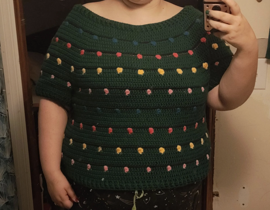 Person wearing a crocheted sweater with colorful yarn dots, taking a mirror selfie