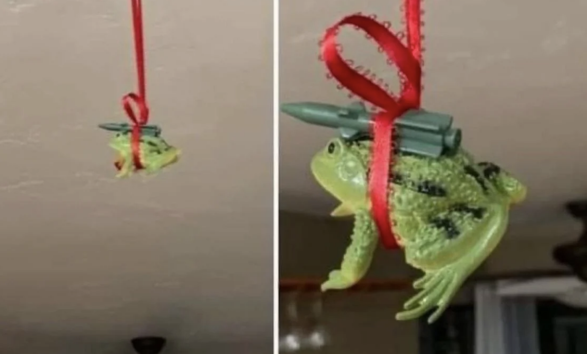 Ornament of a frog tied with a red ribbon, carrying toy missiles on its back, hanging from a ceiling