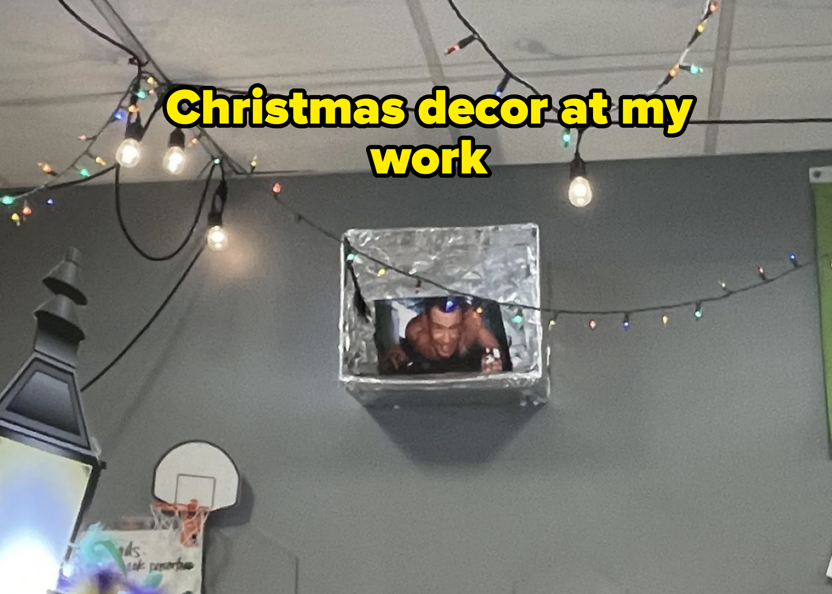 Man in a small metallic box appears to be popping out through ceiling; he&#x27;s playfully gesturing, surrounded by string lights and part of a basketball hoop visible