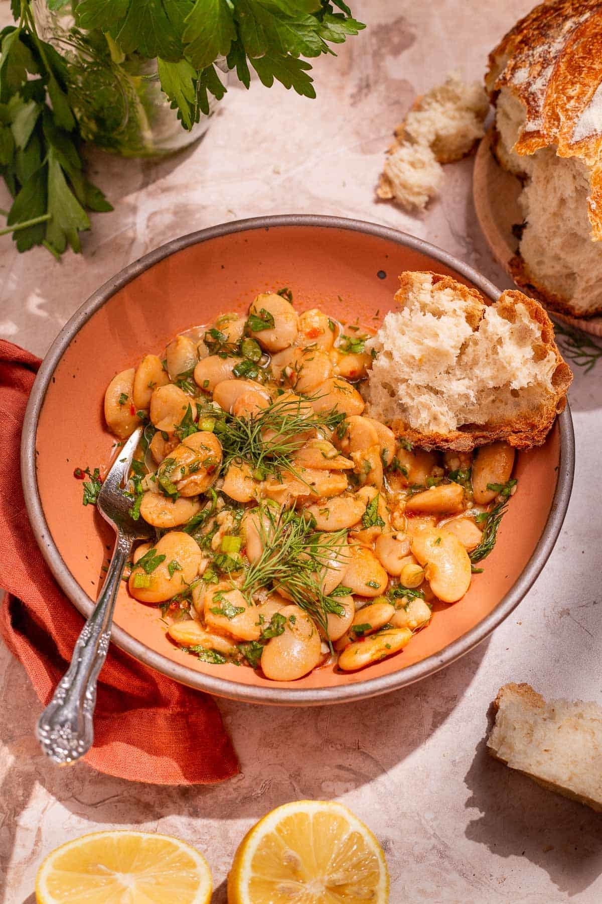 A bowl of seasoned lima beans garnished with herbs, served with a chunk of bread and a cut lemon nearby