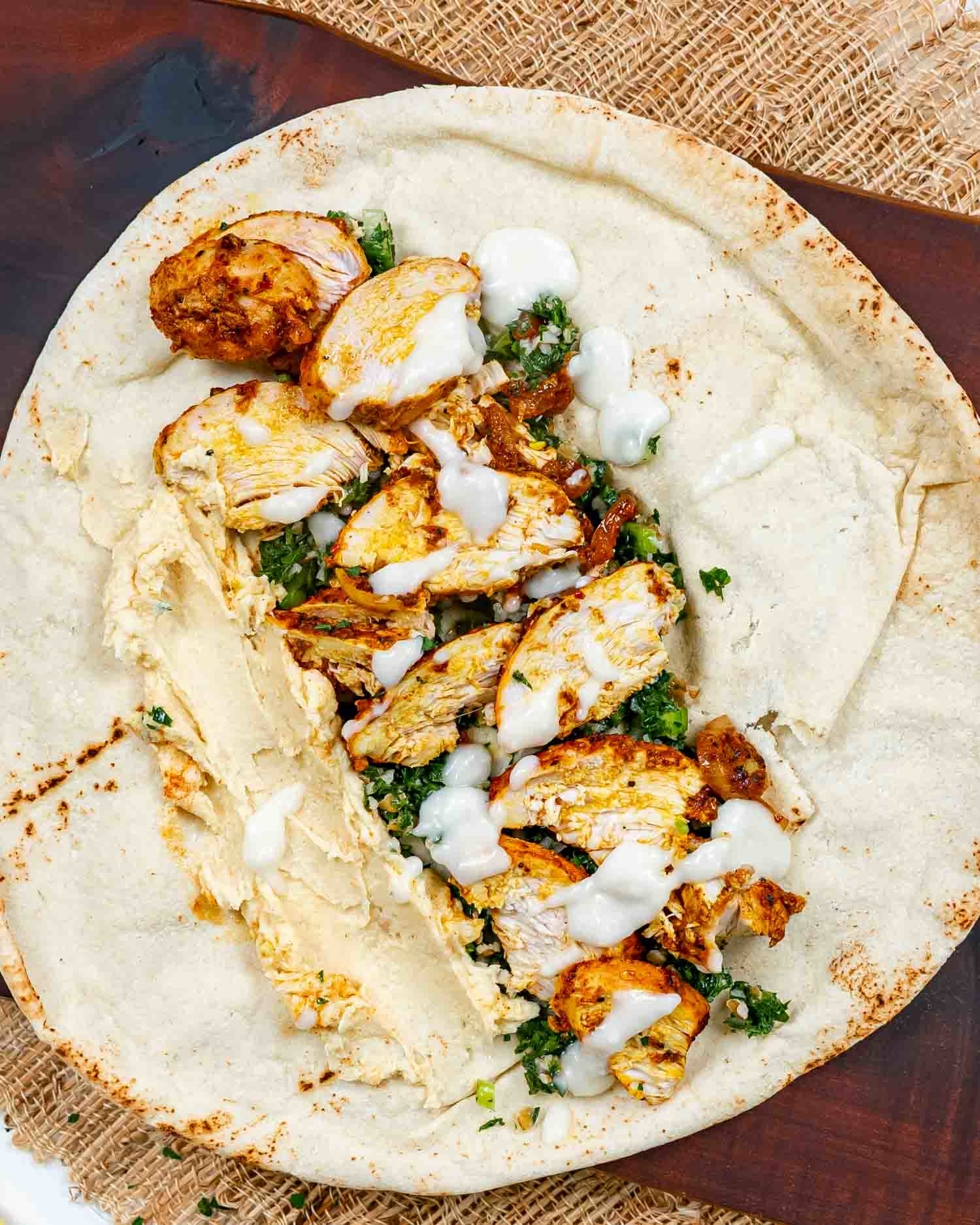 Grilled chicken wrap with hummus, tabbouleh, and a drizzle of white sauce on flatbread, served on a wooden board with burlap background