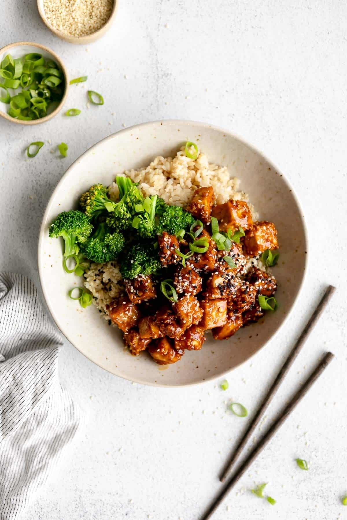 A bowl of sesame chicken served with rice, broccoli, and green onions, accompanied by chopsticks and small bowls of sesame seeds and scallions nearby