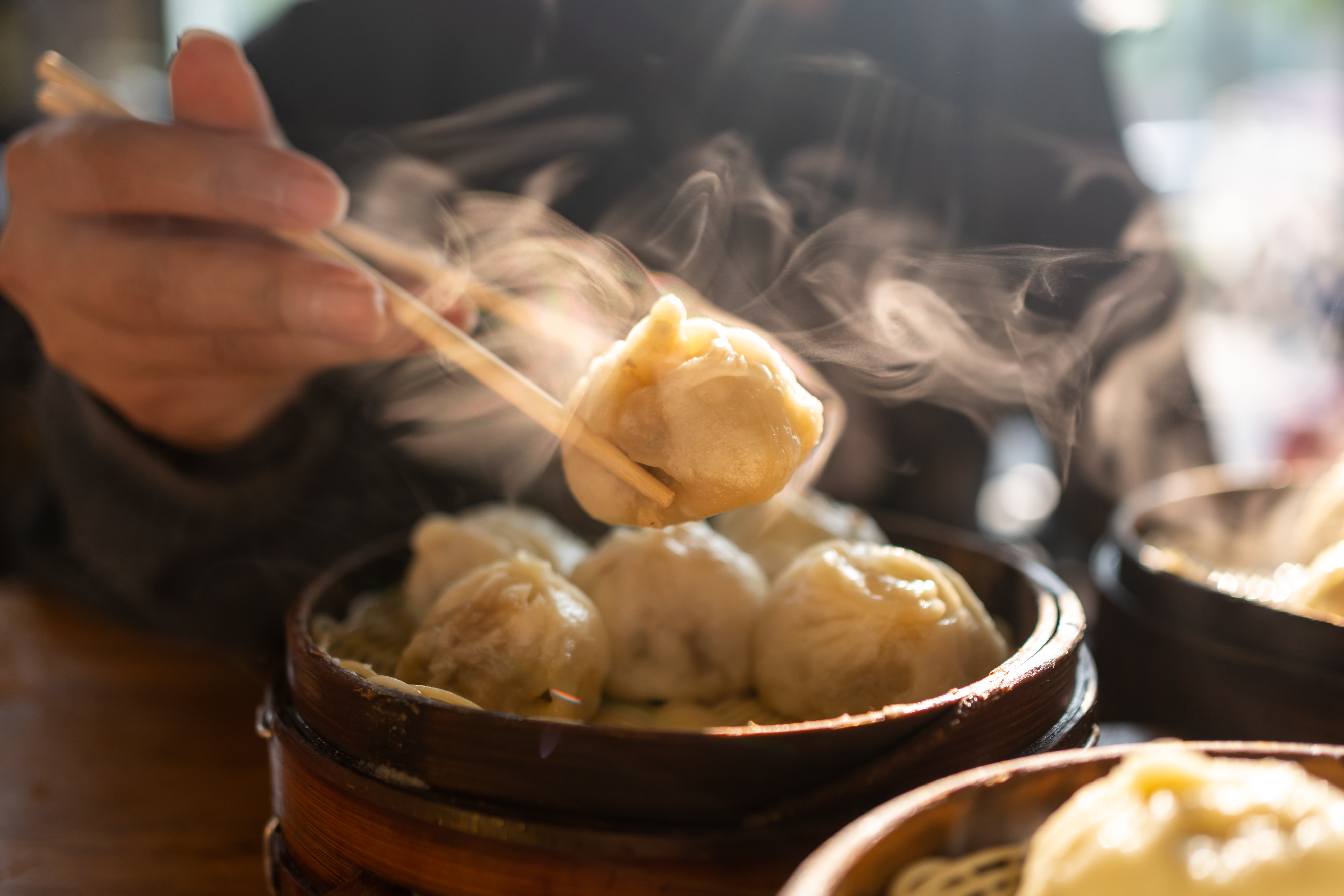 Steaming dumplings in a bamboo basket; a hand uses chopsticks to pick one up.