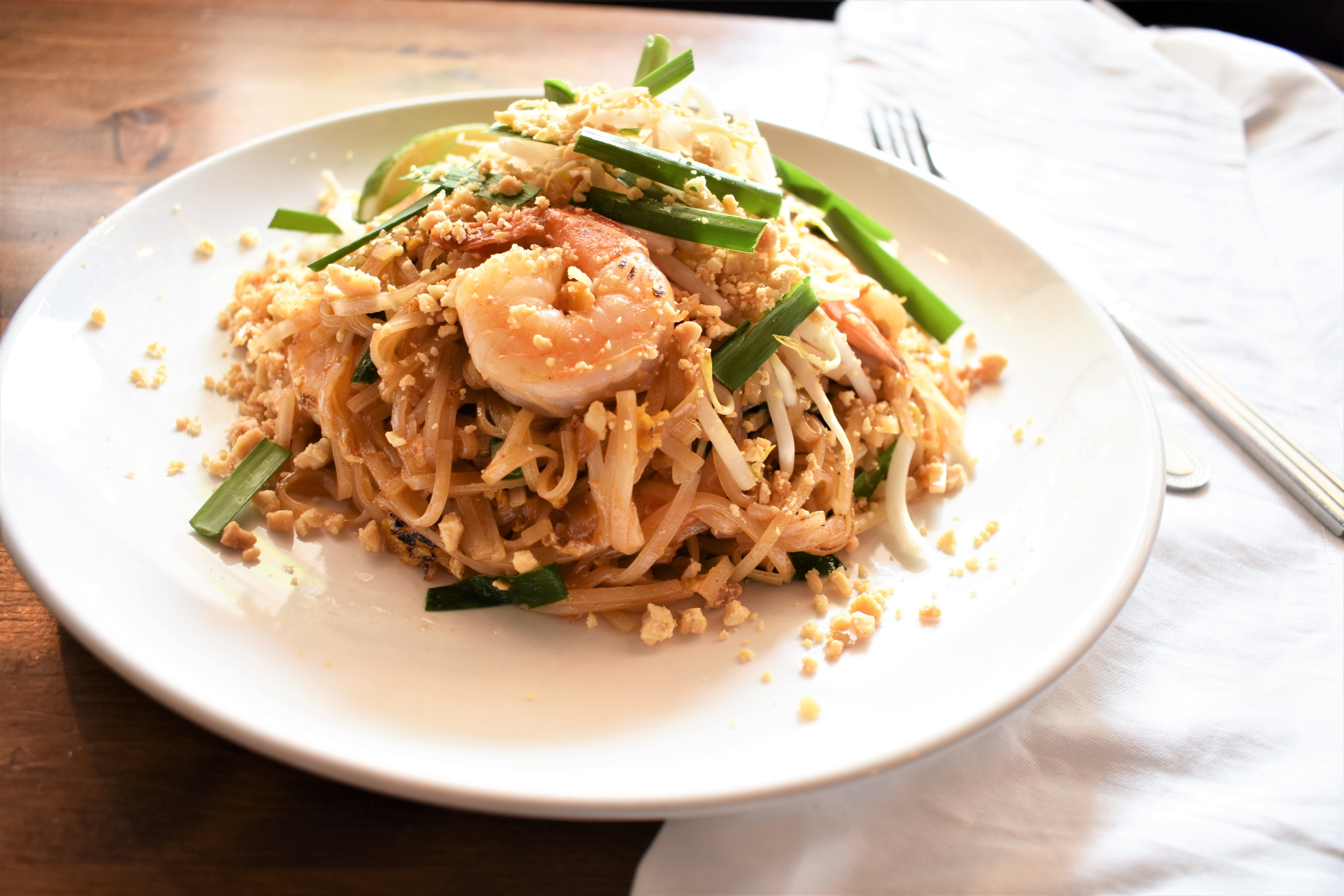 A plate of shrimp pad thai garnished with crushed peanuts and green onions on a wooden table.