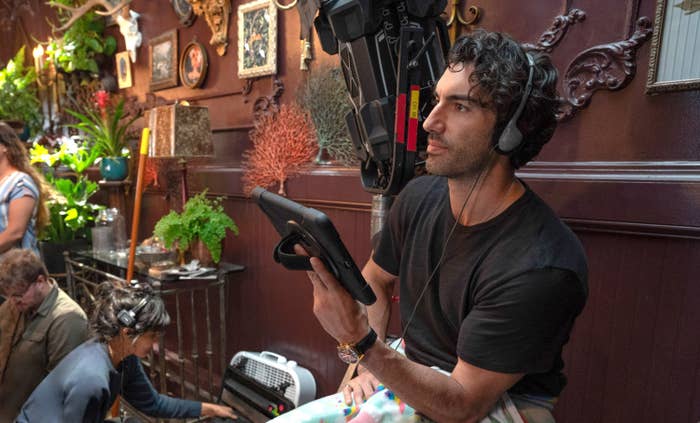 Justin Baldoni on a film set holds a tablet and wears headphones. Crew members work in the background amidst various decorations
