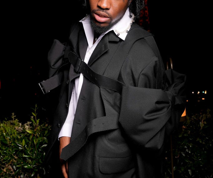 Person wearing a stylish, avant-garde black suit with oversized details on a dimly lit background