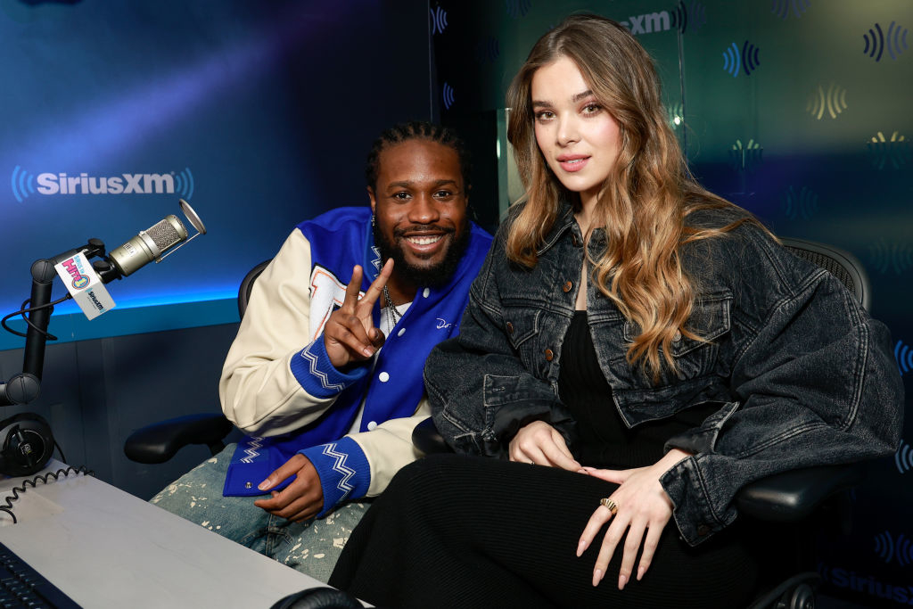 Two individuals at a radio station. One wears a varsity jacket, making a peace sign; the other is in a denim jacket. They sit by a microphone