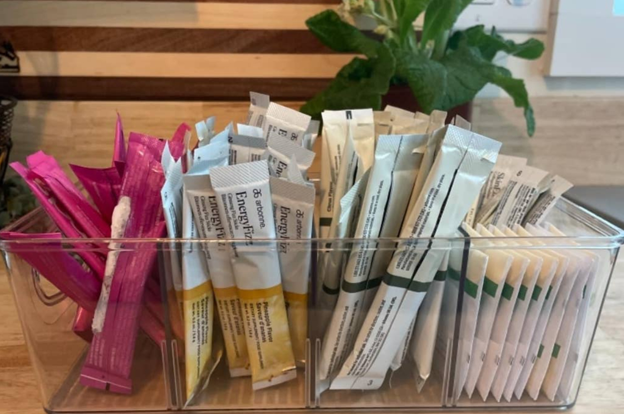 Organized packets of various drink mixes are stored in a clear container on a kitchen countertop, with plants in the background