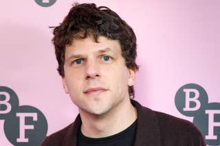 Jesse Eisenberg on red carpet wearing a dark jacket over a black shirt, standing in front of a graphic background