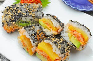 おいしそうな寿司ロールが盛り付けられた白い皿に乗っている。隣に少量のキムチと青い小皿が置かれている。