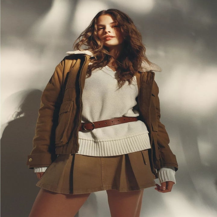 Person in a cozy outfit: shearling-lined jacket, sweater, pleated skirt, and belt, posed in a shopping editorial setting