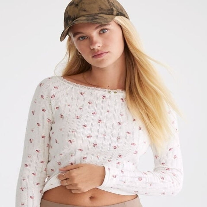 Person in a white floral sweater and cap poses, showcasing casual fashion for a shopping article