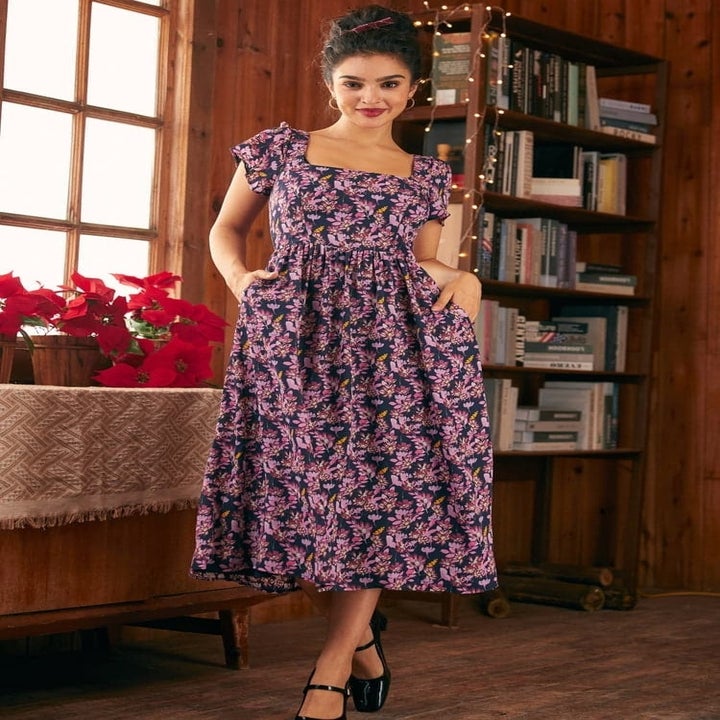 A woman in a floral dress with flutter sleeves stands in a cozy room with bookshelves and flowers
