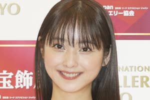 女性が黒いドレスを着て笑顔で立っています。背景にイベントの看板が見えます。