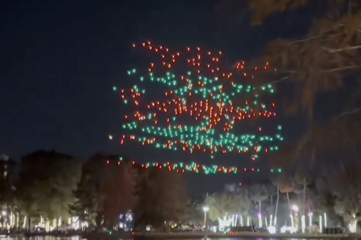 ドローンが夜空に赤と緑で形成した図形が浮かんでいる風景。地面には木々と光が点在している。