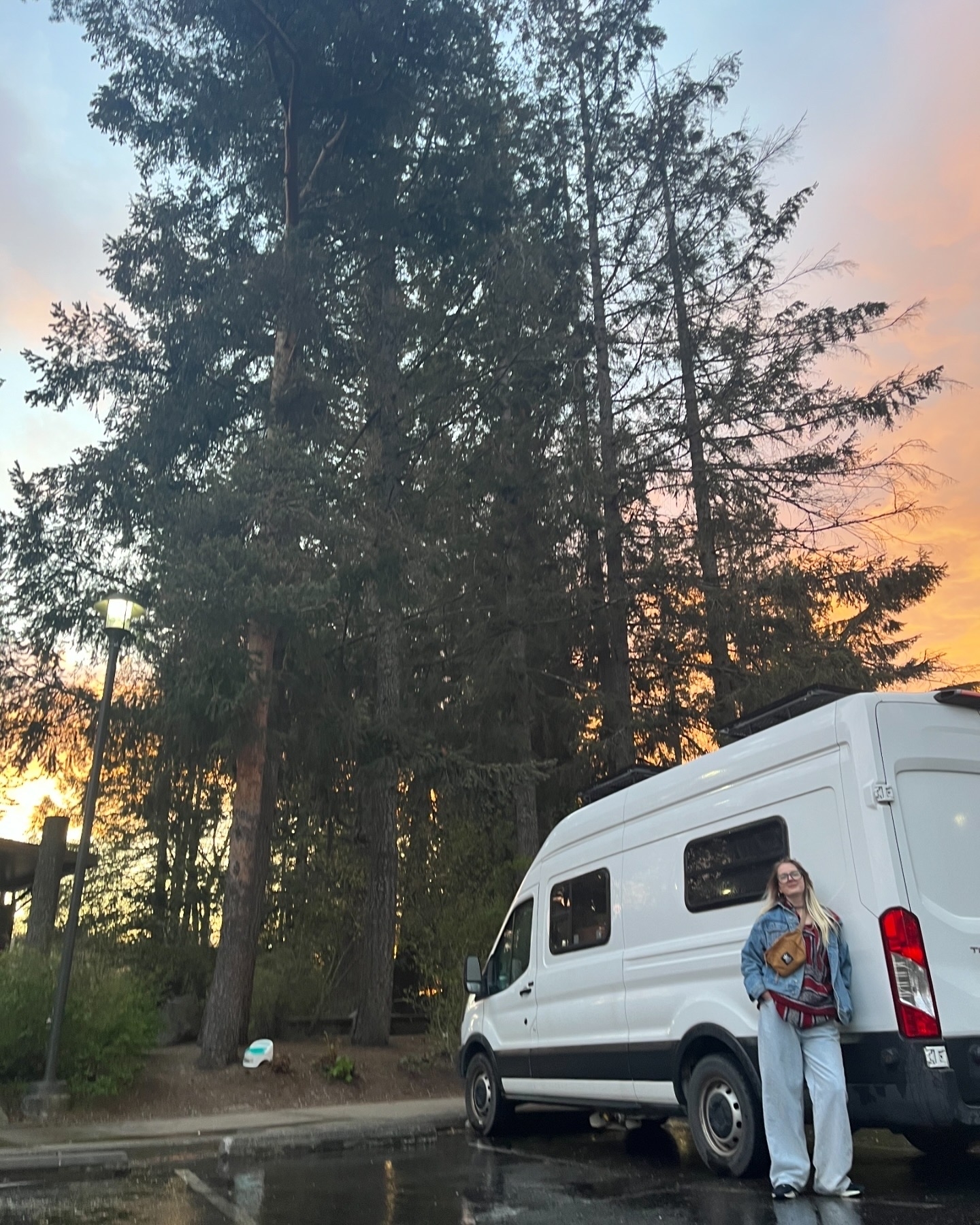 Person stands near a camper van in a parking lot, surrounded by tall trees at sunset. They are wearing casual clothing and a denim jacket