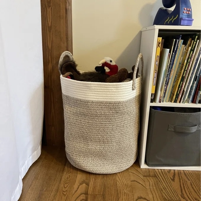 Woven storage basket filled with plush toys next to a small bookshelf and a fabric bin on a wooden floor